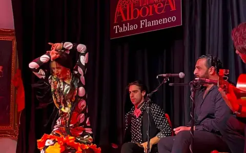Una bailaora de flamenco, con un vestido de lunares y volantes, actúa en un tablao. A su lado, músicos la acompañan con guitarra y cante.