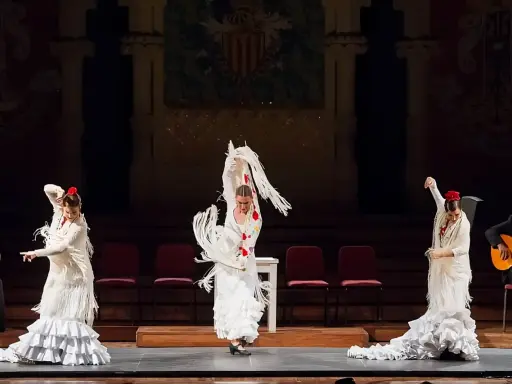 Tres bailarinas de flamenco, con trajes blancos y volantes, ejecutan un **taconeo flamenco** en un escenario. Un guitarrista acompaña la actuación.