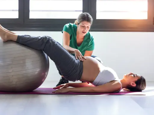 Entrenadora ayuda a una embarazada a hacer ejercicios con pelota en el gym.