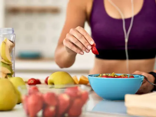 Mujer en ropa deportiva prepara su desayuno saludable con frutas frescas, pensando en qué desayunar antes de entrenar.