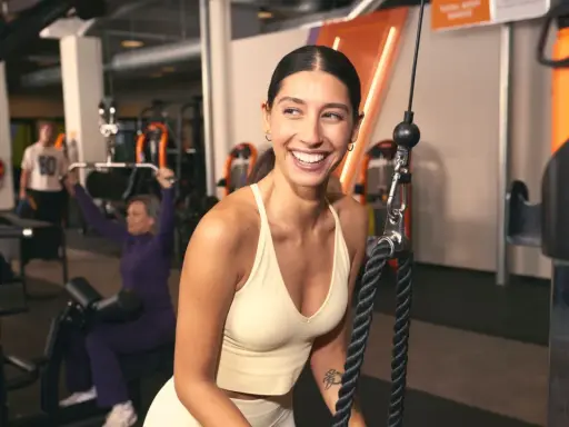 Mujer sonríe en gimnasio, mientras otros realizan ejercicios compuestos y aislados en máquinas.