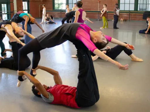 Un grupo de bailarines practica una coreografía compleja, con un hombre en el suelo sosteniendo a una mujer en el aire, evocando la pasión de un flamenco.