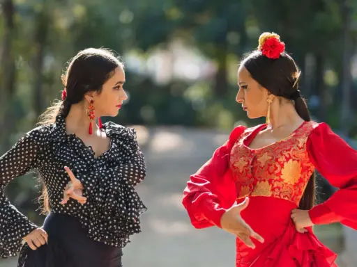 Dos mujeres practican baile flamenco para principiantes en un parque, una con traje de lunares y otra de rojo vibrante.