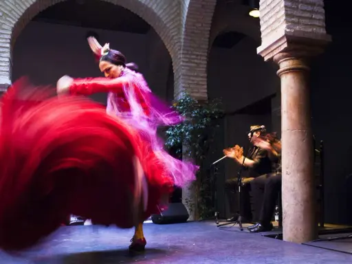 Una bailaora española gira con su vestido rojo, creando un torbellino de tela. A su lado, un guitarrista acompaña con su música.