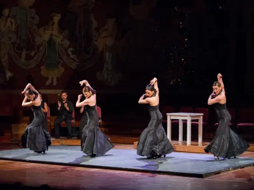 Cuatro bailarinas de flamenco, vestidas de negro, ejecutan pasos apasionados. Un percusionista las acompaña.