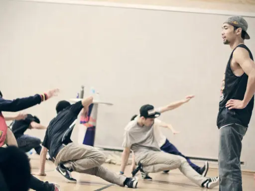 Jóvenes practicando una coreografía de break dance, con movimientos en el suelo y un instructor observando.