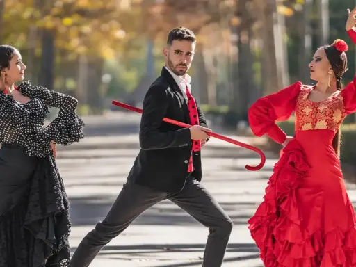 Bailarines de flamenco, uno con traje negro y rojo, sostiene un bastón rojo, mientras dos mujeres con trajes vibrantes ejecutan pasos. Explora los tipos de palos flamencos.