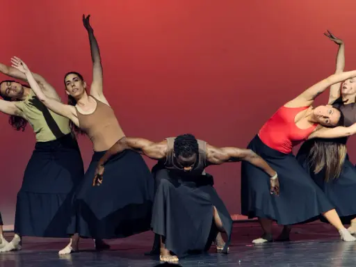 Un grupo de bailarines realiza movimientos fluidos en unas clases de danza, con faldas negras y un fondo rojo vibrante.