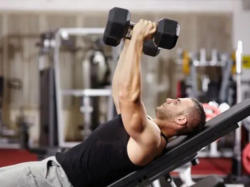 Hombre en gimnasio haciendo press de banca con mancuernas. Un ejercicio clave en una tabla de entrenamiento para ganar masa muscular y perder grasa.