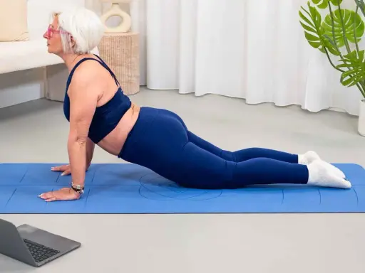 Mujer mayor con pelo blanco realiza postura de cobra en una esterilla azul, parte de su entrenamiento para mujeres.