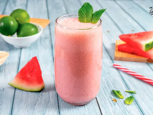 Refrescante zumo de sandía en vaso alto, decorado con menta. Alrededor, trozos de sandía, limones y una pajita a rayas.