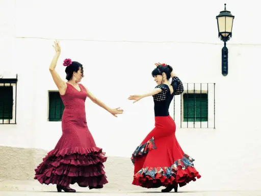 Dos mujeres con trajes vibrantes ejecutan apasionados bailes flamencos frente a una pared blanca.