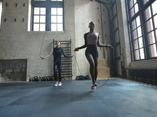 Dos mujeres realizan ejercicios de calentamiento general saltando la cuerda en un gimnasio industrial.