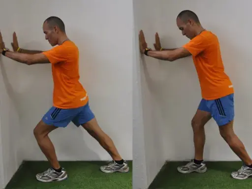 Hombre realizando estiramientos después de correr contra una pared, apoyando las manos y extendiendo una pierna hacia atrás para estirar el gemelo.