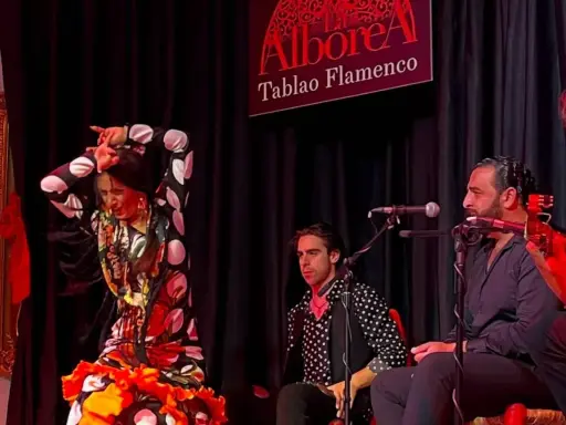 Una bailaora de flamenco, con un vestido de lunares y volantes, actúa en un tablao. A su lado, músicos la acompañan con guitarra y cante.