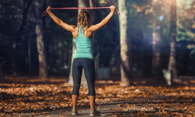 Mujer entrenando tren superior con tabla de ejercicios y gomas elásticas en un parque otoñal.