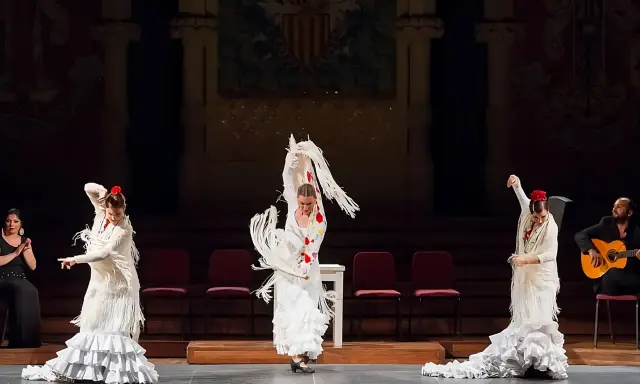 Tres bailarinas de flamenco, con trajes blancos y volantes, ejecutan un **taconeo flamenco** en un escenario. Un guitarrista acompaña la actuación.