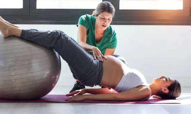 Entrenadora ayuda a una embarazada a hacer ejercicios con pelota en el gym.