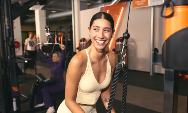 Mujer sonríe en gimnasio, mientras otros realizan ejercicios compuestos y aislados en máquinas.