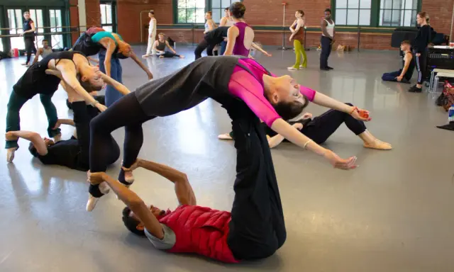 Un grupo de bailarines practica una coreografía compleja, con un hombre en el suelo sosteniendo a una mujer en el aire, evocando la pasión de un flamenco.