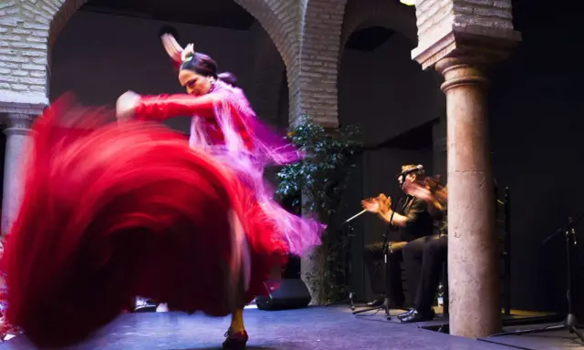 Una bailaora española gira con su vestido rojo, creando un torbellino de tela. A su lado, un guitarrista acompaña con su música.