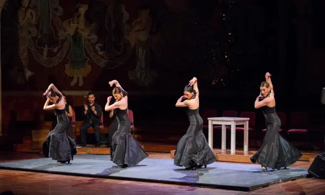 Cuatro bailarinas de flamenco, vestidas de negro, ejecutan pasos apasionados. Un percusionista las acompaña.