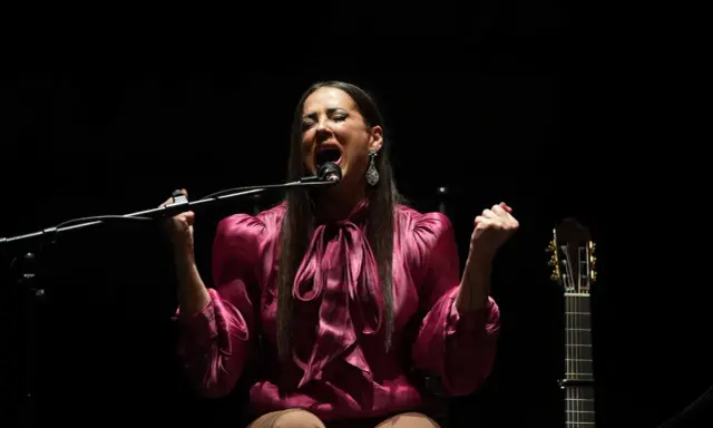 Cantante flamenco mujer, con el alma desbordada, canta apasionadamente frente al micrófono. A su lado, una guitarra espera.