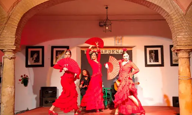 Tres bailarinas de flamenco, vestidas de rojo y con abanicos, ejecutan fandangos flamencos en un escenario.