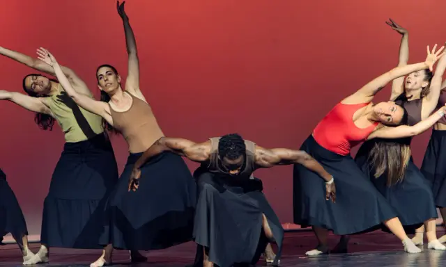 Un grupo de bailarines realiza movimientos fluidos en unas clases de danza, con faldas negras y un fondo rojo vibrante.