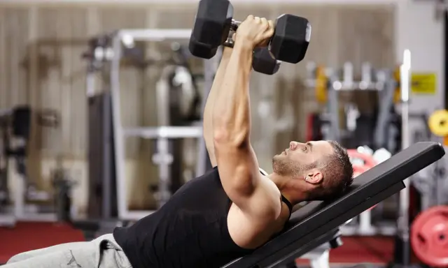Hombre en gimnasio haciendo press de banca con mancuernas. Un ejercicio clave en una tabla de entrenamiento para ganar masa muscular y perder grasa.