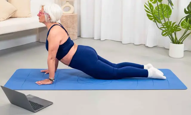 Mujer mayor con pelo blanco realiza postura de cobra en una esterilla azul, parte de su entrenamiento para mujeres.