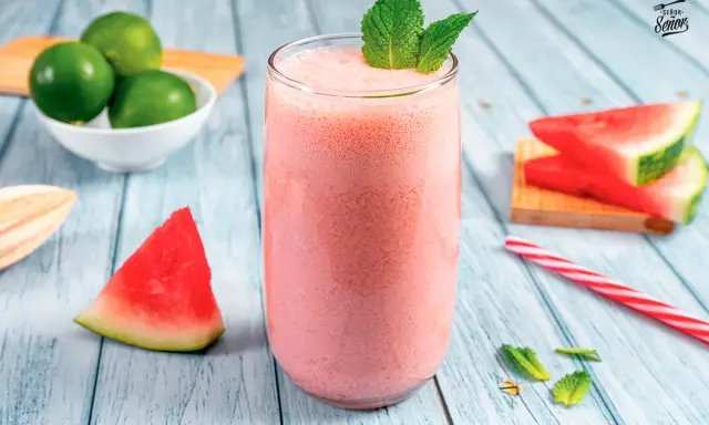 Refrescante zumo de sandía en vaso alto, decorado con menta. Alrededor, trozos de sandía, limones y una pajita a rayas.