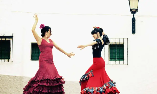 Dos mujeres con trajes vibrantes ejecutan apasionados bailes flamencos frente a una pared blanca.