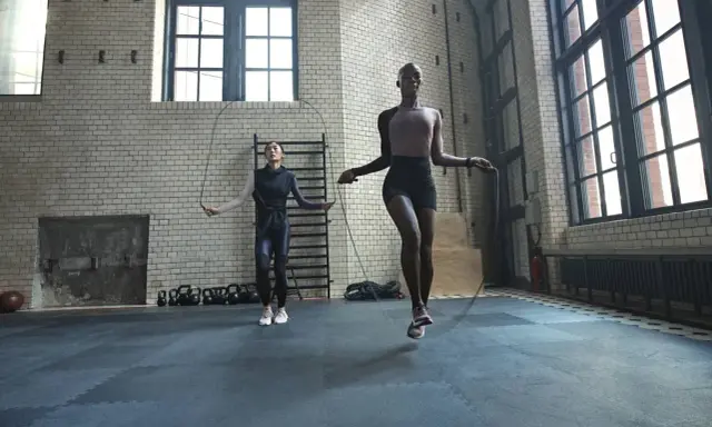 Dos mujeres realizan ejercicios de calentamiento general saltando la cuerda en un gimnasio industrial.