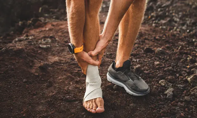 Corredor con tobillo vendado, sintiendo dolor espinilla al correr. Reloj deportivo naranja en muñeca.
