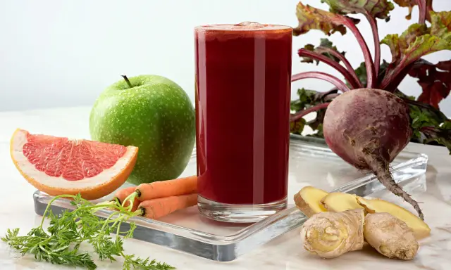 Vaso de zumo de remolacha vibrante rodeado de ingredientes frescos: remolacha, zanahorias, manzana verde, pomelo y jengibre.
