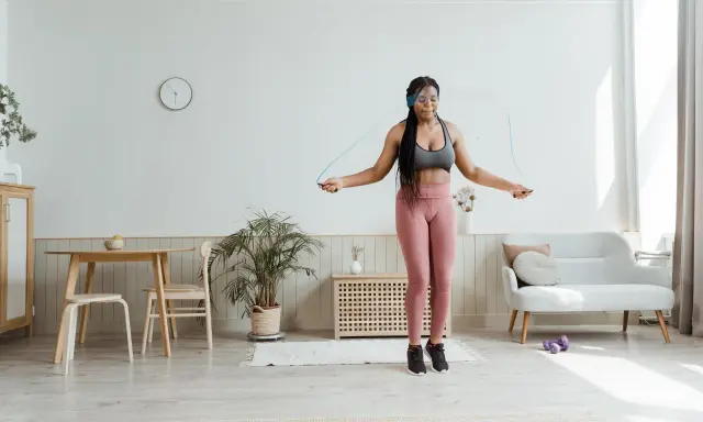 Mujer joven con auriculares realiza ejercicio hit saltando la cuerda en su sala de estar.