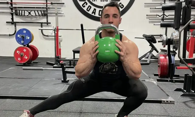 Hombre realiza desplantes laterales con pesa rusa verde en un gimnasio.