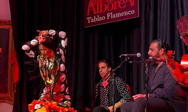 Una bailaora de flamenco, con un vestido de lunares y volantes, actúa en un tablao. A su lado, músicos la acompañan con guitarra y cante.