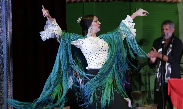 Bailarina de flamenco con mantón verde vibrante, sus movimientos evocan frases flamenco. Un hombre aplaude al fondo.