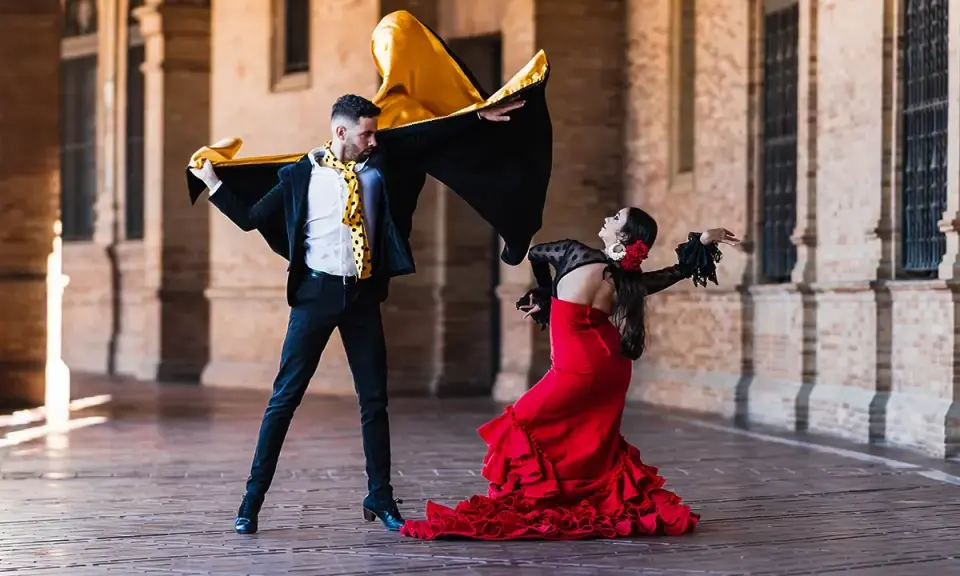 Pareja bailando flamenco con pasión. Él, con capa negra y dorada, ella, con vestido rojo voluminoso. Un jaleo flamenco lleno de arte.