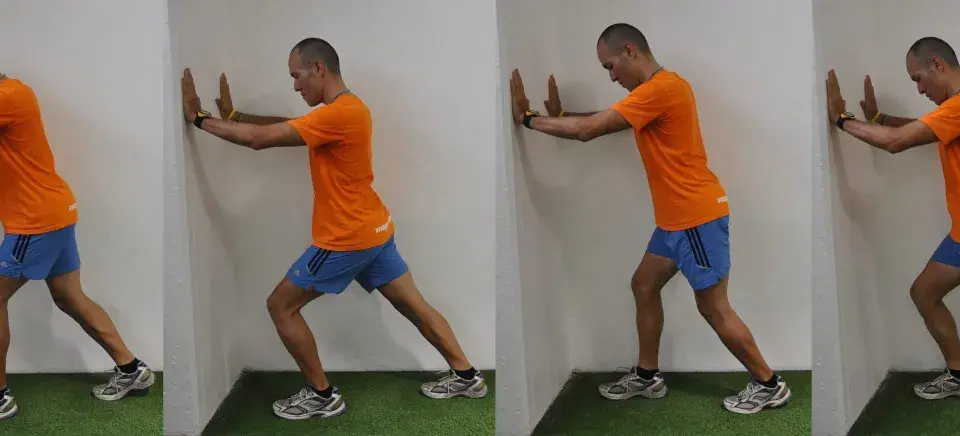 Hombre realizando estiramientos después de correr contra una pared, apoyando las manos y extendiendo una pierna hacia atrás para estirar el gemelo.