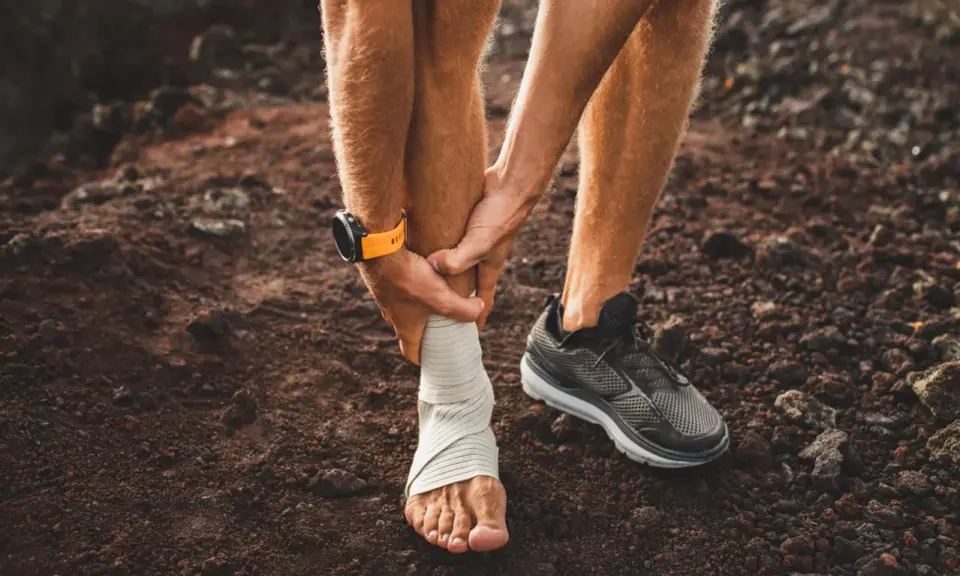 Corredor con tobillo vendado, sintiendo dolor espinilla al correr. Reloj deportivo naranja en muñeca.