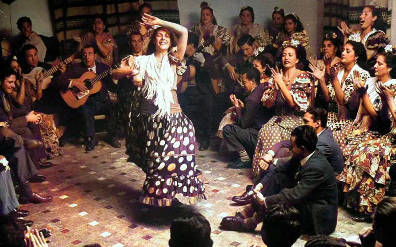 Una mujer baila flamenco con un vestido de lunares, rodeada de m&uacute;sicos y espectadores aplaudiendo. La zambra es un espect&aacute;culo vibrante.