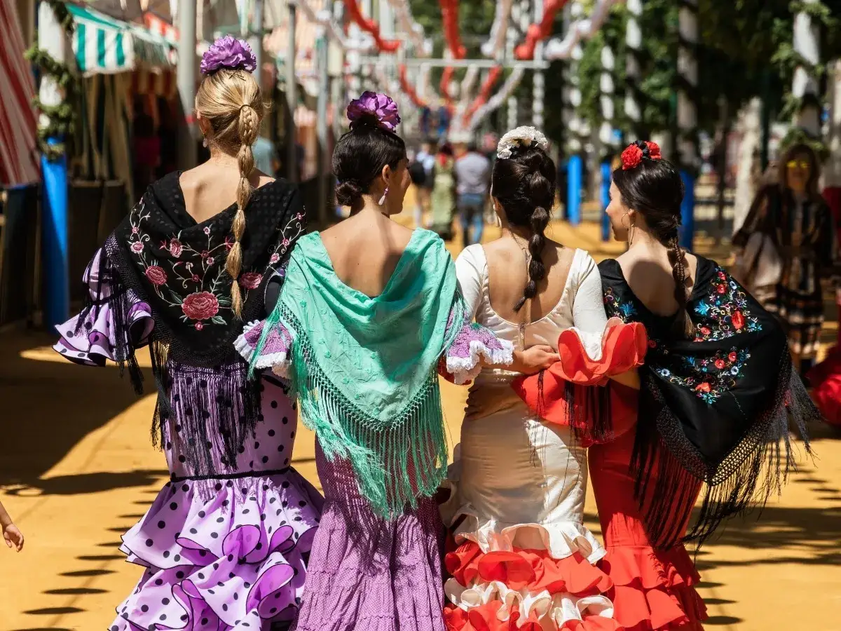 Cuatro mujeres lucen distintos tipos de trajes de flamenca, con volantes, mantones bordados y flores en el pelo, paseando en un día soleado.