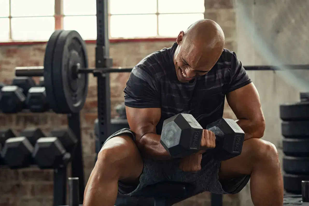 Hombre calvo entrenando bíceps con mancuernas, con expresión de esfuerzo para estar fuerte.