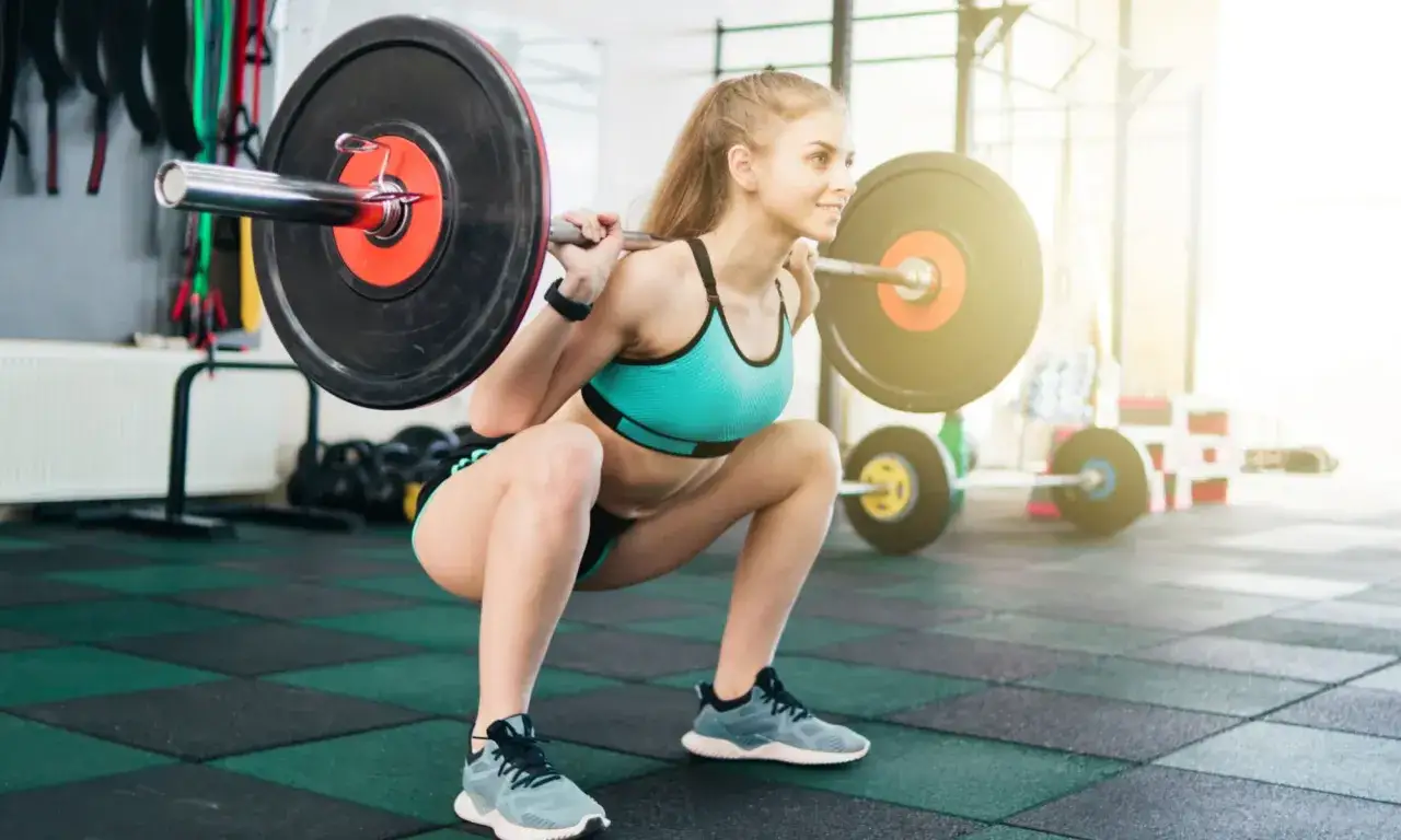 Mujer joven realiza sentadillas con barra, un ejercicio compuesto. En el gimnasio, se entrena con ejercicios compuestos y aislados.