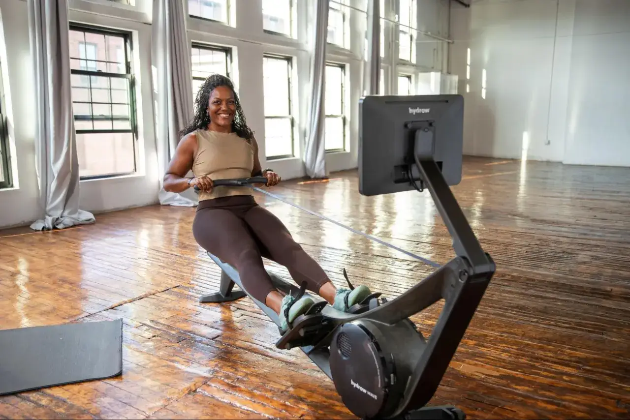 Mujer sonriente entrenando en m&aacute;quina de remo, disfrutando de una sesi&oacute;n de cardio hiit en un estudio luminoso.
