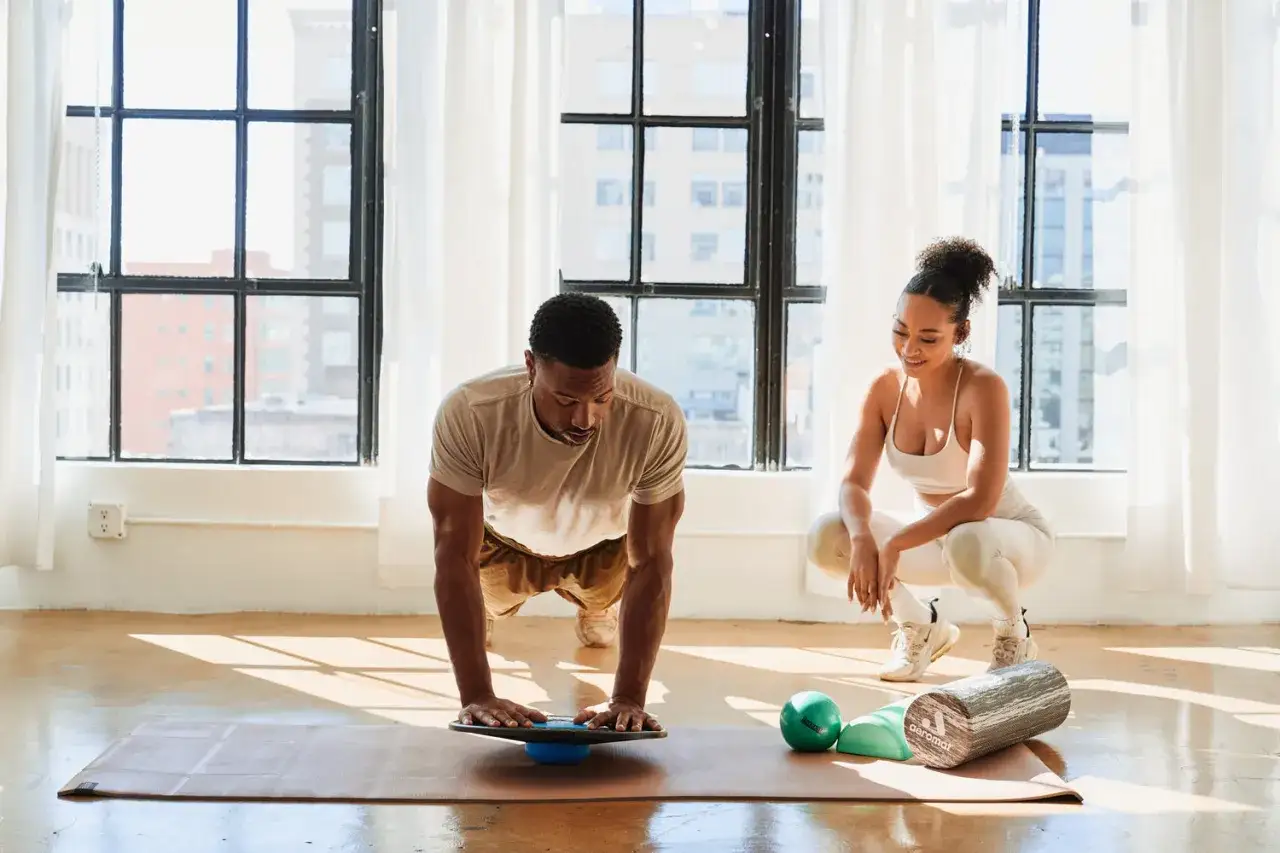 Pareja entrenando en casa para perder peso. Él hace plancha sobre una tabla de equilibrio, ella lo observa sonriendo.