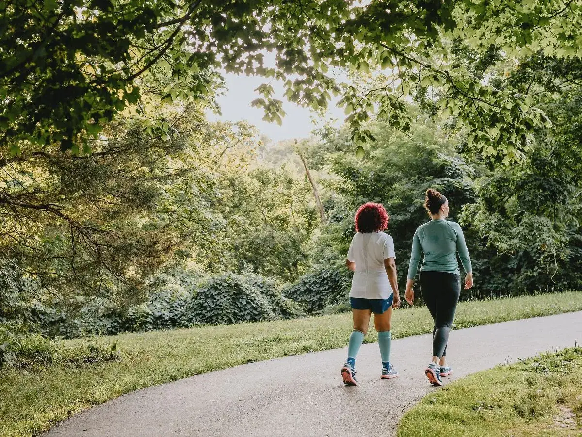 Dos mujeres disfrutan de un paseo por un sendero arbolado, alternando entre correr y andar a intervalos.